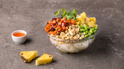 Poke bowl with rice, salmon, edamame beans, parsley, pineapple and peanuts on a gray textured background. Next to the sweet chili sauce and pineapple slices. Copy space. Close up
