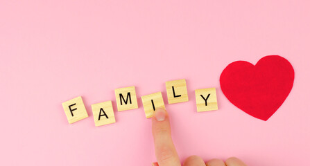 The word family from wooden letters and a red heart on a pink background. Female hand collects the...