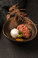In a brown plate, beef tartare with capers, white sauce, micro greenery and spices. Dark textured background, black napkin, bronze decorative twig. Close up.
