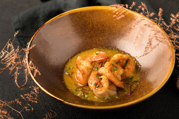 In a bronze-colored plate, a dish of grilled shrimp in a sauce with herbs. Black textured background, black napkin, decorative bronze twig. Close up.