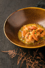 In a bronze-colored plate, a dish of grilled shrimp in a sauce with herbs. Black textured background, black napkin, decorative bronze twig. Close up.