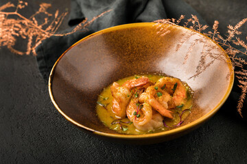 In a bronze-colored plate, a dish of grilled shrimp in a sauce with herbs. Black textured background, black napkin, decorative bronze twig. Close up.