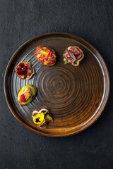 Round brown plate with 5 tapas with beef roast beef, tartare, salmon, chicken and avocado. A dish of haute cuisine. Dark gray textured background. Top view.