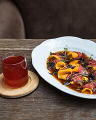 Oval plate with beef carpaccio with prunes, wine, sweet potatoes and micro greenery. On a wooden table in a restaurant, a transparent mug with red tea.