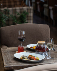 On the table in the restaurant serving for 2 persons. An oval dish with fish and eggplant, a glass teapot with red tea, a mug of tea, a glass of  wine, a plate with carpaccio in the background.