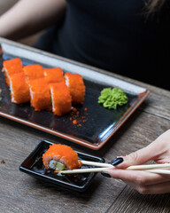 A girl holds a sushi roll with chopsticks over soy sauce. On the wooden table in front of her is a rectangular plate with sushi, wasabi and ginger. Close up