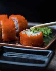 Sushi rolls on a rectangular plate close-up. Chopsticks put wasabi on the roll. In the foreground is soy sauce in a black sauce pan. Close up.
