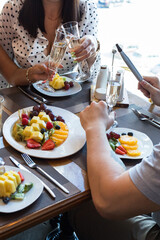 2 girls and 1 guy at a table in a restaurant clink glasses of champagne. The table is served with fruit and berries plates.