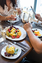 2 girls and 1 guy at a table in a restaurant clink glasses of champagne. The table is served with fruit and berries plates.