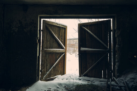 View From The Inside Of An Abandoned Garage, Shabby And Dirty Concrete Walls, Thick Wooden Gates Ajar And A Snow-covered Space Is Visible Behind Them, Nature Always Takes Its Toll