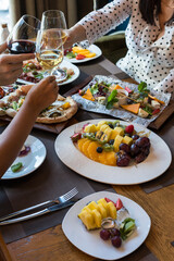 3 hands with glasses of champagne clink glasses over a table with snacks, a fruit plate, a set of sushi rolls, a cheese plate. Top view