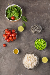 On a gray textured background, a bowl with spinach leaves, a bowl with cherry tomatoes, a bowl with rice, a bowl with edamame beans and sesame seeds. Grey textured background. Top view.