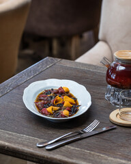 Oval plate with beef carpaccio with prunes, wine, sweet potatoes and micro greenery. On a wooden table in a restaurant, a transparent teapot with red tea, a transparent mug.