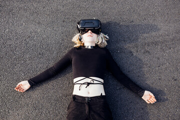 Young nonconformist woman lying floor outdoors using augmented reality 3D headset