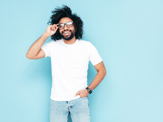 Portrait of handsome smiling hipster  model.Unshaven Arabian man dressed in white summer t-shirt and jeans clothes. Fashion male with long curly hairstyle posing near blue wall in studio. Isolated