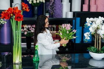 Fototapeta premium A young female florist takes care of flowers in a cozy flower shop and collects bouquets. Floristry and making of buckets in a flower shop. Small business.