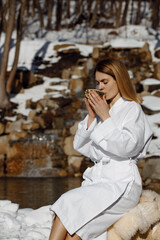 Beautiful young model after spa drinking hot tea on the street against the backdrop of winter forest, wearing a white hotel robe