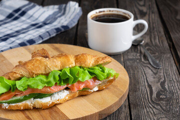 breakfast sandwich, croissant with red fish, salmon, cottage cheese, cucumbers and salad on a dark wooden background with coffee and napkin.