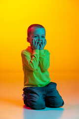Studio shot of cute little boy, kid sitting on floor isolated on yellow studio backgroud in neon light. Concept of child emotions, facial expression, childhood