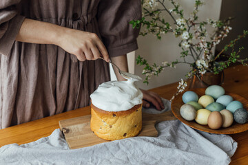 Woman decorating homemade easter bread with sugar glaze on rustic table with natural dyed eggs and spring blossom in room. Happy Easter! Woman baking stylish easter cake
