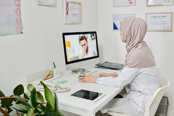 Young Muslim female doctor in hijab consulting online male patient with covid coughing or sneezing in front of computer screen