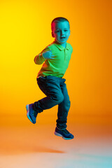 Fototapeta na wymiar Full-length portrait of charming little boy posing isolated on yellow studio backgroud in neon light. Concept of child emotions, facial expression, action, childhood