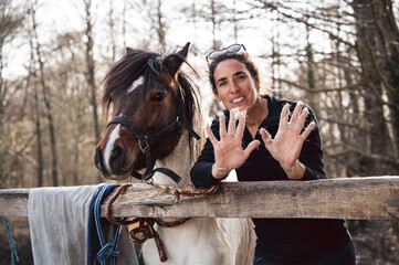 Beautiful smile woman standing next to her cute pony and showing hands full with horse hair in spring shedding season on countryside ranch. Horse molt shower.
