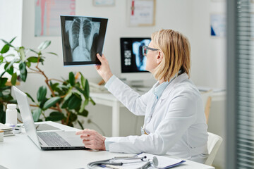 Blond female radiologist in whitecoat looking at x-ray image of patient lungs while sitting in...