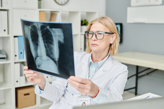 Serious mature radiologist in eyeglasses looking at x-ray image of lungs of patient while sitting by workplace in medical office