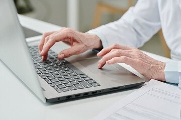 Fototapeta na wymiar Hands of mature female clinician pressing buttons of laptop keyboard while sitting by desk and consulting patients online