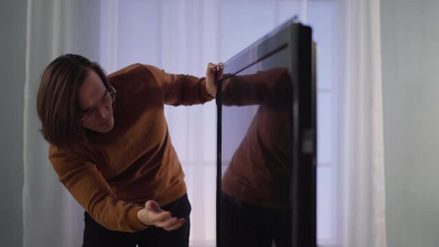 Confused man slams smart TV screen with trenches against window curtains in apartment slow motion. Hipster and broken TV set at home. Damaged appliance