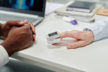 Hand of mature female patient with pulse oximeter on fingertip sitting in front of doctor during check-up of oxygen level in blood