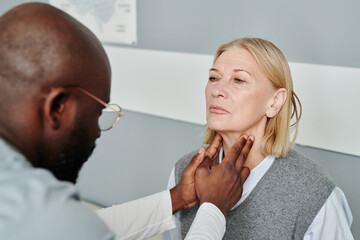 Mature blond female patient having checkup of her thyroid glands by African American male...