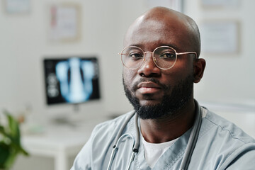 Young serious African American male general practitioner in blue uniform and eyeglasses sitting by workplace in front of camera in hospital - obrazy, fototapety, plakaty