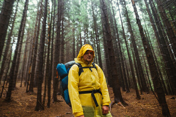 Portrait of a male tourist in a yellow raincoat standing in a wet rainforest on a hike and looking at the camera with a serious face.