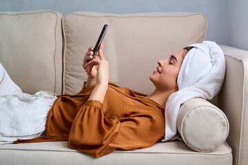 Side view of young happy female with smartphone relaxing on beige sofa with white towel on her head...