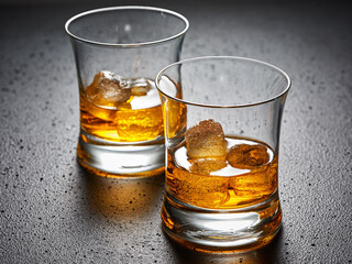 Two glasses of elegant whiskey with ice on black textured background.