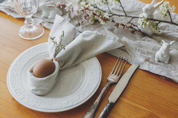 Stylish Easter brunch table setting. Easter egg in bunny napkin on plate with cutlery, bunny, spring flowers and rustic cloth on wooden table. Easter table arrangement and eco friendly decorations