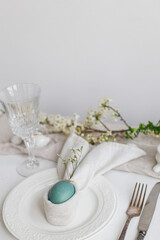 Stylish elegant Easter brunch table setting. Easter egg in bunny napkin on plate with cutlery, bunny, spring flowers on rustic white table. Modern Easter table arrangement