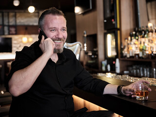 Portrait of an attractive happy man holding a glass of whiskey and talking on his mobile phone at the bar or pub.