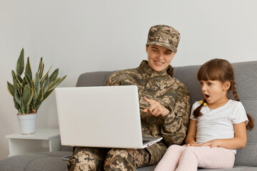 Portrait of happy delighted military woman wearing camouflage uniform and cap and her impressed daughter, looking at notebook screen with amazement.