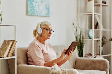 Side view portrait of modern senior woman using tablet and headphones while sitting on sofa in comfy home interior, copy space