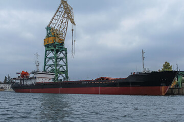A dry cargo ship stands in the port for loading or unloading. In the background is a port crane and a cloudy sky.