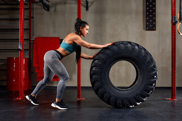 Fototapeta na wymiar Fit female athlete working out with a huge tire, turning and flipping in the gym. Woman exercising with big tire.