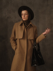 A beautiful young woman poses in the studio on a gray background. Vintage look. Autumn or spring coat, black hat and scarf. Large eye arrows.