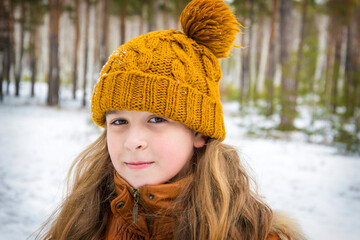 In winter, a beautiful girl with long hair in a hat with a balabon stands in the forest.