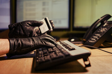 A man in black gloves with a plastic credit card in his hands at a computer keyboard in a dark...