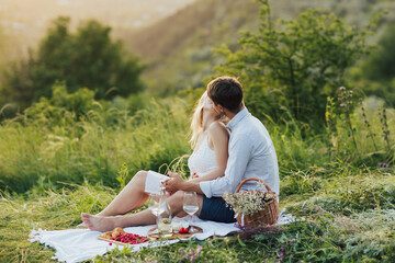 A loving couple sitting on a date on a picnic in the park. Young man and woman sit on a blanket with wine and fruits near and kissing.