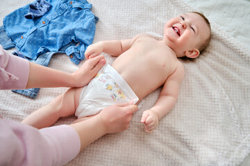 A mother woman puts on a diaper for a baby boy. Mom dresses a happy child in clothes on the bed.
