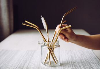 Child hand take golden metal drinking straw from glass jar in home kitchen. Sustainable lifestyle concept.